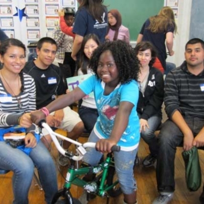 Five college students, sitting down on chairs, pose for a picture with a young girl sitting on bicycle wearing a helmet. In the background are more young children and college students.