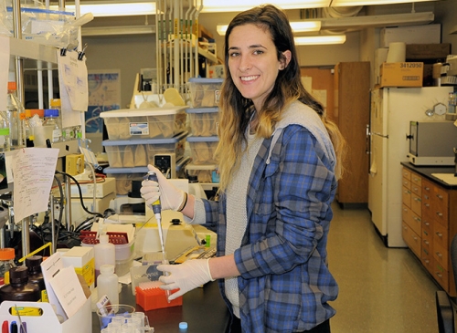 Female student in a lab flannel wearing shirt and latex gloves smiles for camera.