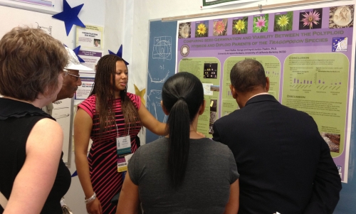 Student in classroom presents her STEM research poster to a small group of students and faculty.