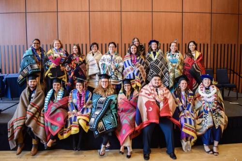 18 people wearing different colorful Native American blankets are smiling at the camera. Some students are wearing lei and graduation caps.