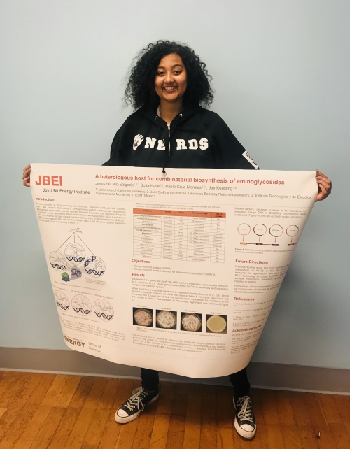 A student in a black Cal NERDS hoodie is smiling and holding a research poster.