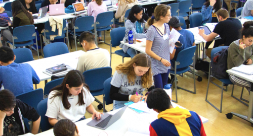 Many students are seated in long white tables with blue chairs at the Student Learning Center. Some students work independently, while others work in groups. Some students are using laptops, while others are writing on paper. A female tutor wearing glasses and a nametag walks down with a paper and phone on her left hand.