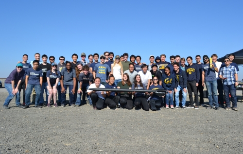 A group of forty-three individuals standing in rows outdoors smiling for a group photo. The first row of individuals is holding a model rocket.