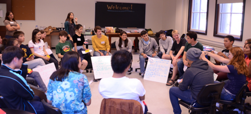 Students are sitting at the center of the classroom in a circle, smiling and discussing something. Two of the students in the center are holding large white sheets with some writing on them. The blackboard in the background reads "welcome".
