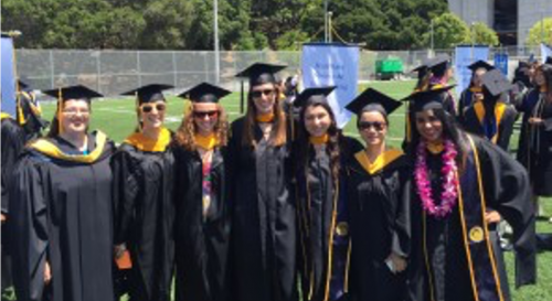 A group of recent graduates, wearing their full cap and gown, pose for a picture on a football field.