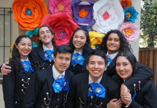 Several members of the mariachi in uniform pose for a picture with a large floral decoration as a backdrop.