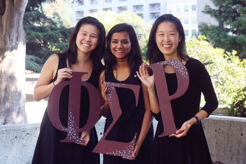 Three students are smiling as they hold the letters of their sorority in their hands.