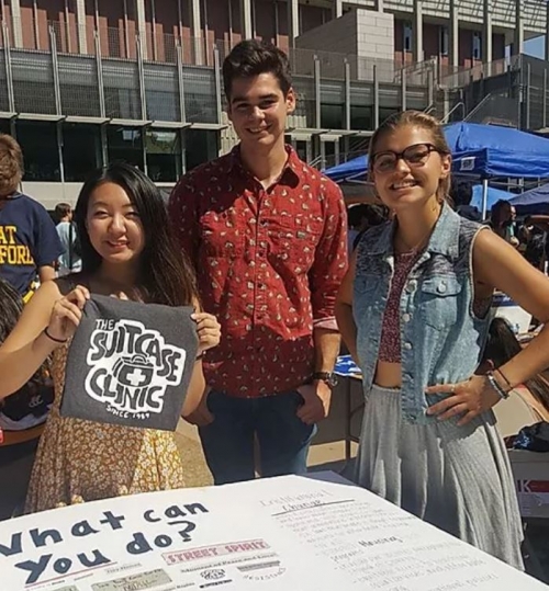 Three students smiling and standing in front of a "What can you do poster" at an outdoor tabling event. One student holds a folded tshirt with program logo.