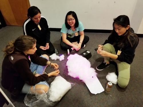 Group of four female students sit on the floor inside a room. The students each wear a nametag. There is a big bag of cotton, an object creating pink light, and an aerosol can on the floor.