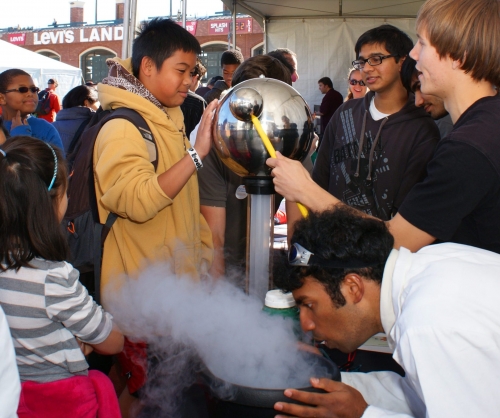 A group of college students performs science experiments in front of kids who visited their booth. A young male touches a metallic sphere as a college student gently taps it with a smaller metallic sphere. Another college student gently blows at steam coming out of a bowl.