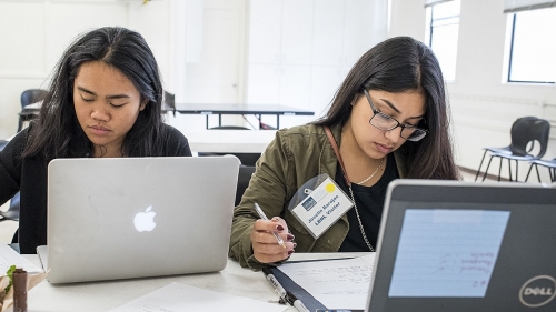 Two female students are sitting in front of their computers in an empty classroom. One of them is writing something down on the piece of paper, the other reads something from her piece of paper while holding a pen.