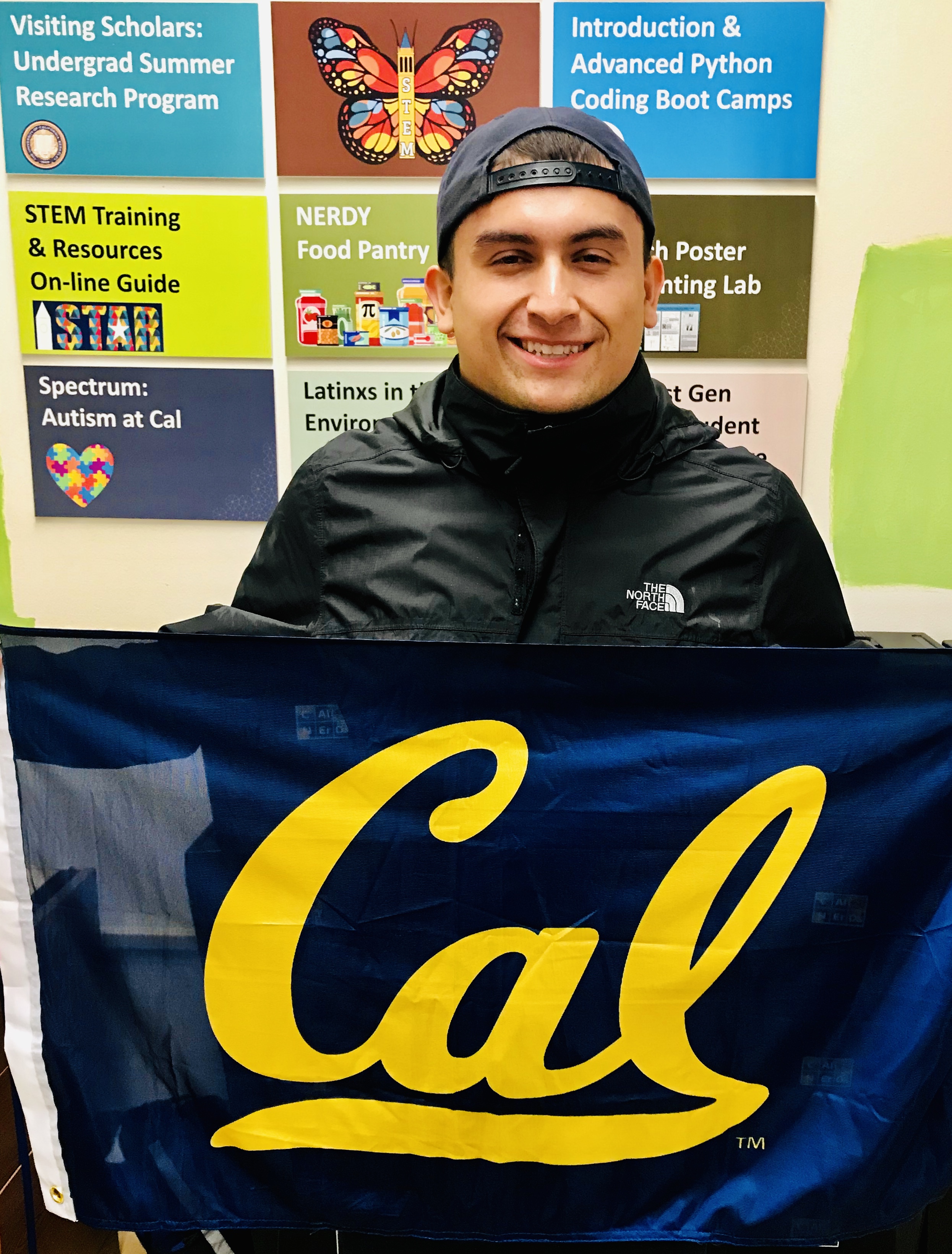 A young man stands in front of science-related posters holding a flag that reads 'Cal' and smiling