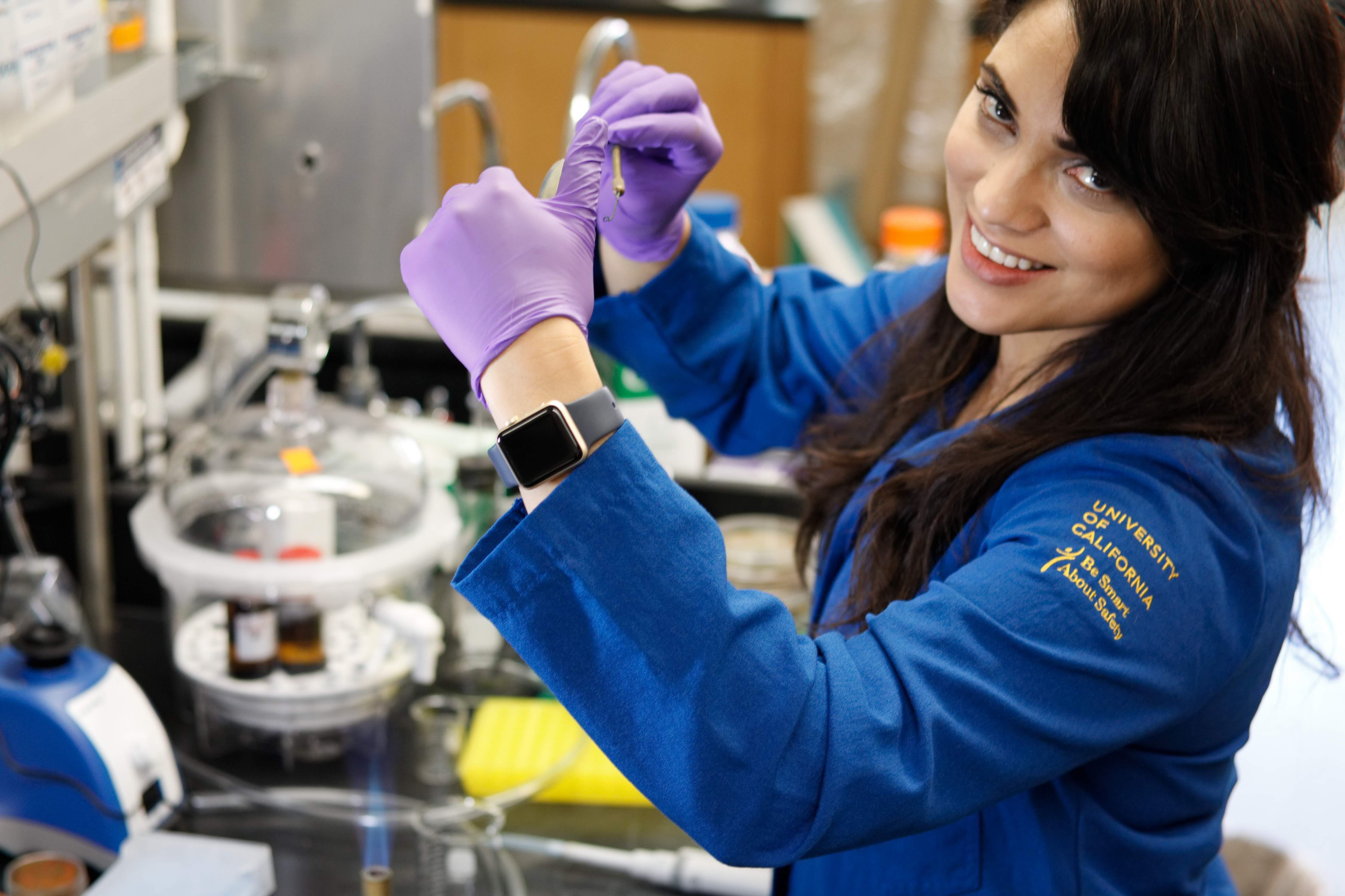 Smiling woman scientist working in lab wearing laboratory coat, latex gloves, and an Apple watch.