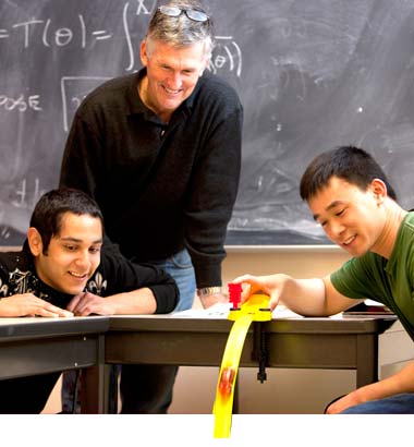 A group of three men at a desk in front of a classroom chalkboard viewing what seems to be a demonstration.