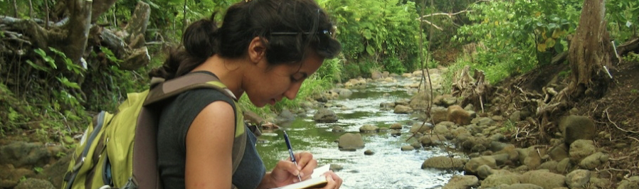 A female student wearing a backpack and hair in a ponytail, writes in a notepad while standing in a small forest stream.