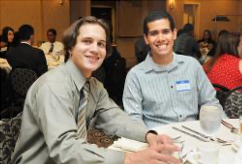 Two male students are sitting at the dining table smiling at the camera. More people of different ages and genders are sitting at the other tables in the background.
