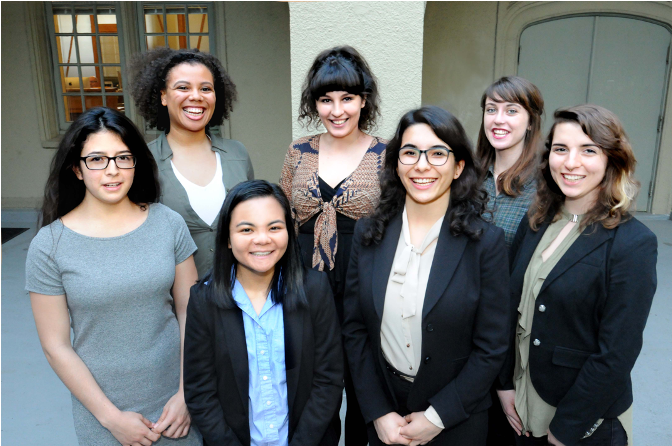 Seven females in professional clothes are smiling at the camera at the light-gray patio of the campus building.
