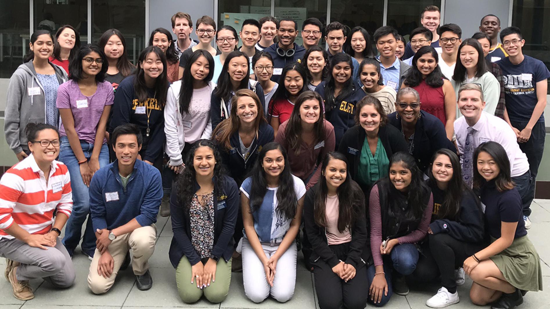 A group of 44 computer science scholars huddled together and smiling for a group photo outside of UC Berkeley's computer science building