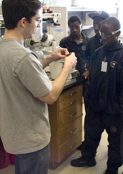 A young man in a grey shirt giving a demonstration to three chemistry scholars who are all in matching black uniform.