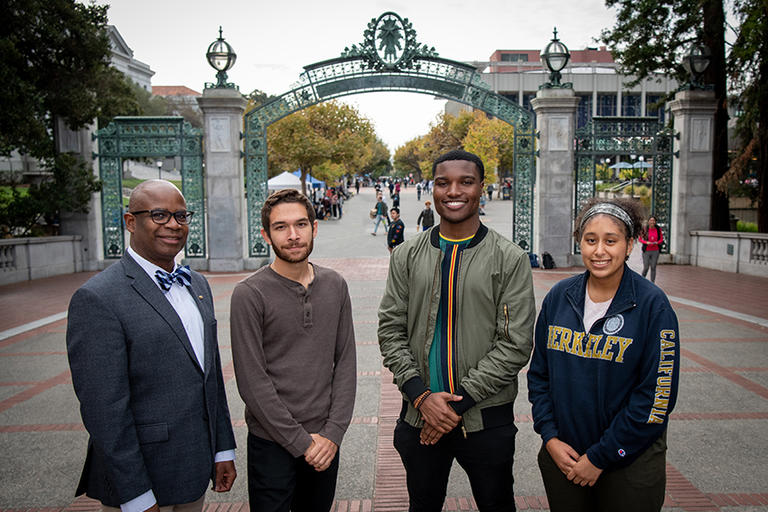 Three students pose next to SEED Scholar Program Director, Ira Young, in front of the Sather Gate. All four individuals are smiling.