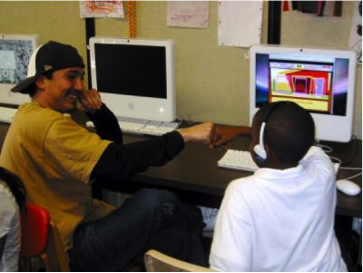 Older male student smiles towards, and fist-bumps, younger male student. Both are sitting down in front of computers.