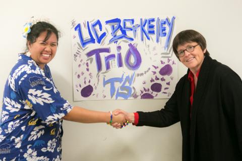 Woman in blue floral print shirt shakes hands with woman in black shirt and glasses in front of program poster.