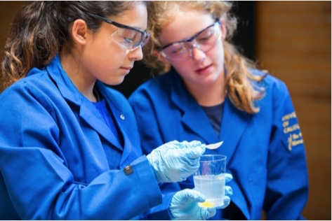 Two young female students in bright blue lab coats and in lab glasses are performing an experiment. one of the students is holding a spoon above a glass with an unidentified light-blue liquid in it, whilst the other student is inclining slightly to observe it.
